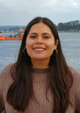 Nicole Delgado-Parada, a young woman with long dark hair, and earrings, wearing a sweater, smiling at the camera.  Behind her is water, a far shoreline with trees, and a boat.