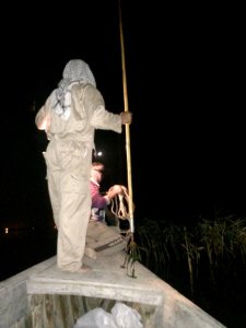 Fisherman at night standing on prow of boat holding long spear with four long prongs.  Click for larger version.