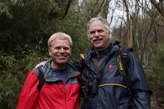 Tom on the left, and Robert ont the right, next to a big tree trunk with woodland behind them.