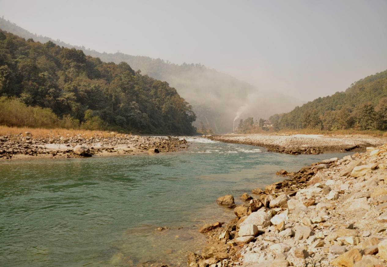 Fork in the river, with wide, barren, rocky bankds leading to dense forest.  THe hill behind is almost obscured by dense smoke issuing from a chimney at a factory on the river bank in the centre of the image. 