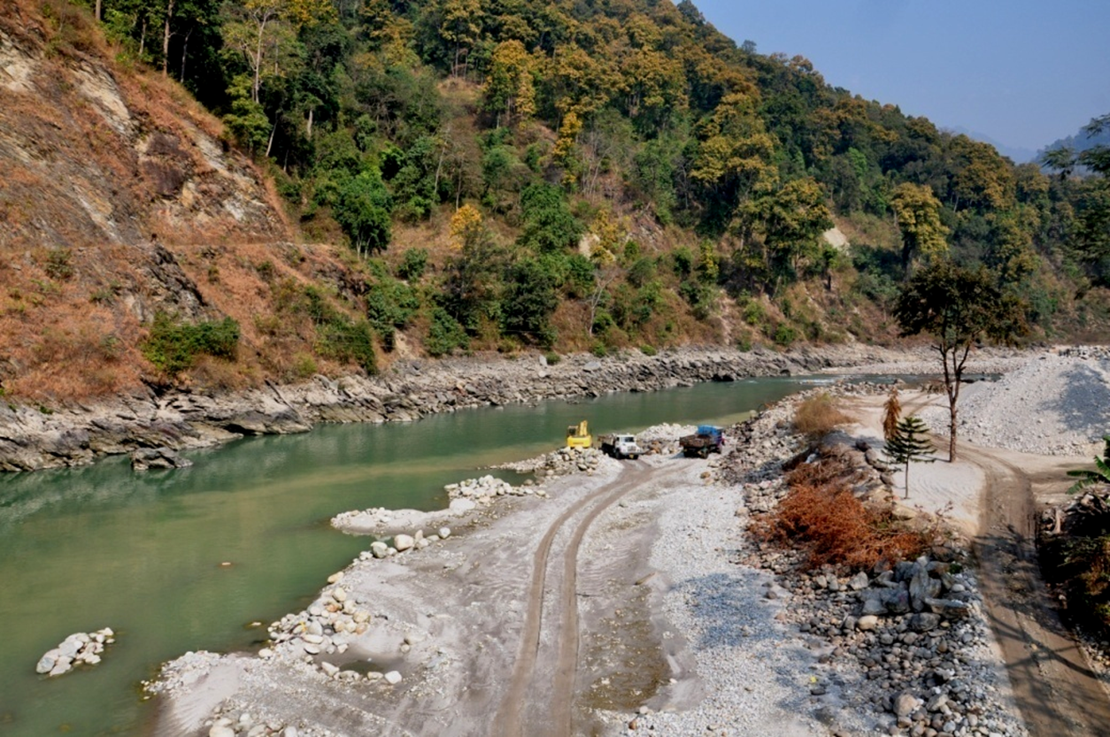 Steep cliff with some vegetation a obove narrow stretch of river where the nearer, shallower bank is being dug away for rock, stone and gravel.  Trucks and tracks and heaps of stone are visible. 